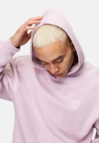 Ljuslila hoodie med en känguruficka, vit textlogotyp på bröstet och huva med dragsko. Mjuk, texturerad tyg.