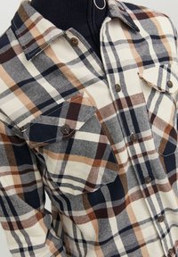 Chemise en flanelle à carreaux dans des tons crème, marine et marron ; dotée de deux poches poitrine et de boutons marron ; texture douce avec un design de col standard.