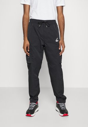 Homme portant un pantalon cargo noir avec un cordon blanc, un t-shirt blanc et des baskets noires avec des détails rouges et blancs, debout sur un fond uni.