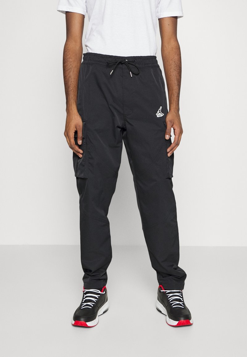 Homme portant un pantalon cargo noir avec un cordon blanc, un t-shirt blanc et des baskets noires avec des détails rouges et blancs, debout sur un fond uni.