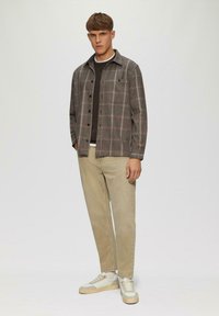 Grau kariertes Hemdjackett über einem dunklen Pullover, kombiniert mit hellbeigen Hosen und weißen Sneakers. Lässiges Design mit Knopfverschluss und Taschen.