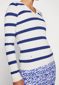 Vit V-ringad tröja med marinblå ränder, ribbad textur och en silveraccent vid fållen. Kombinerad med mönstrade blå byxor.
