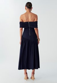 Laivansinis off-the-shoulder mekko, jossa on tiukka yläosa ja valuva, kerroksellinen hame. Selkäpuolella vetoketju, teksturoidusta kankaasta valmistettu ja korostettu vyötärö.