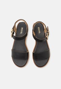Sandalias de cuero negro con dos correas anchas y un cierre de hebilla. Diseño minimalista con una suela marrón y detalles del logo en dorado en la plantilla.