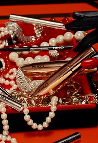 KIKO-Makeupprodukte in einer roten Box angeordnet, darunter eine glanzvolle rosé-goldene Mascara, ein schwarzer Eyeliner und cremefarbene Perlen, mit silbernen Akzenten und Texturen.