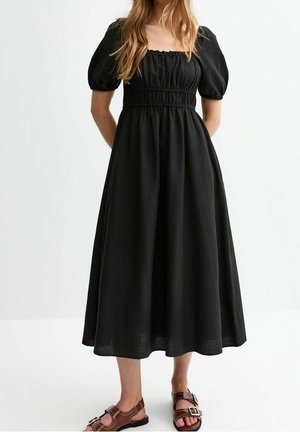 Mujer que lleva un vestido midi negro con mangas abullonadas y escote cuadrado, combinado con sandalias marrones con hebilla, de pie frente a un fondo blanco.