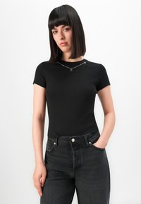 Woman with straight black hair wearing a fitted black short-sleeve top and dark high-waisted jeans, posing with one hand in pocket.