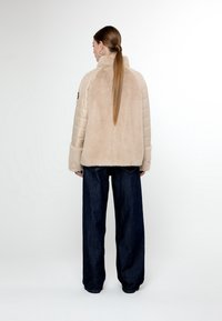Giacca in fur sintetico beige con maniche trapuntate, colletto alto e polsini larghi, abbinata a pantaloni a gamba larga in denim scuro. Vista posteriore mostrata.