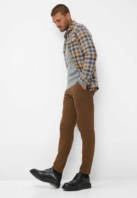 Pantaloni marroni a sigaretta con un maglione grigio sotto una camicia a quadri blu e marrone, abbinati a scarpe nere con lacci.