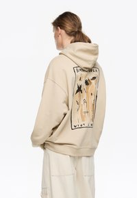 Sudadera beige de gran tamaño con capucha. Presenta un estampado gráfico floral en tonos terrosos en la parte posterior. Material de algodón con un corte holgado.