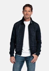 Marineblauwe ritsjack met een doorgestikte voorkant, gebreide mouwen en een staande kraag. Draagt over een wit shirt met jeans, met subtiele textuur.