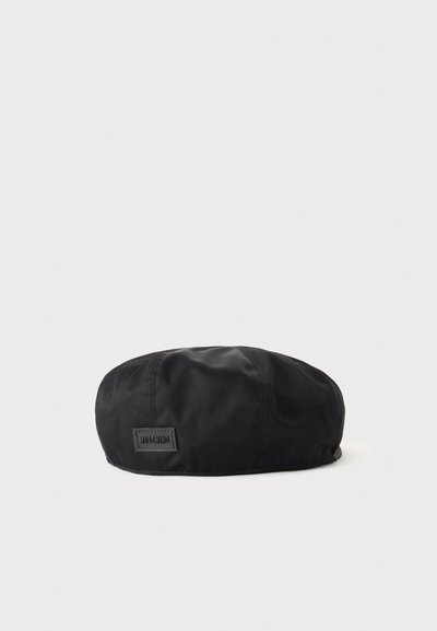 Béret en tissu noir doux avec un petit patch logo Moschino rectangulaire sur le côté, présenté sur un fond clair uni.