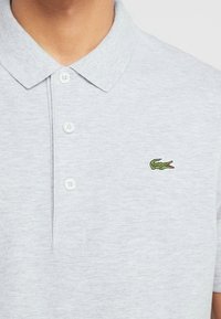 Grå bomullspolo med krage, tre vita knappar och en liten grön broderad krokodil-logotyp på bröstet.