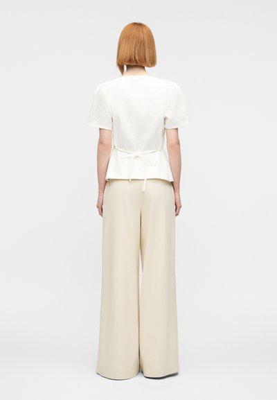 Λευκή λινή μπλούζα με κοντά μανίκια και μπροστινή ζώνη τύπου πεπλούμ, συνδυασμένη με παντελόνια εκρού σε ανοιχτή γραμμή και ψηλή μέση, από λείο ύφασμα.