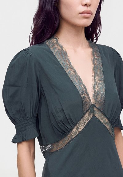 Femme portant une robe vert foncé avec des manches bouffantes et une bordure en dentelle transparente le long du profond décolleté en V et de la taille.