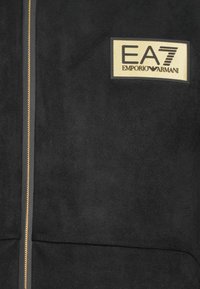 Black zip-up jacket made of soft fabric, featuring a gold and beige EA7 Emporio Armani logo patch and a prominent gold zipper.