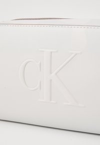 Bílá pouzdro z umělé kůže s embosovaným logem "ck", se zipovým uzávěrem a jemnou lesklou texturou s akcenty šití.