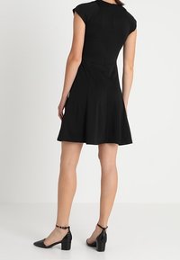 Abito nero aderente con maniche a cappuccio, gonna svasata e dettagli nella cucitura posteriore, abbinato a décolleté nere in pelle verniciata con cinturini alla caviglia.