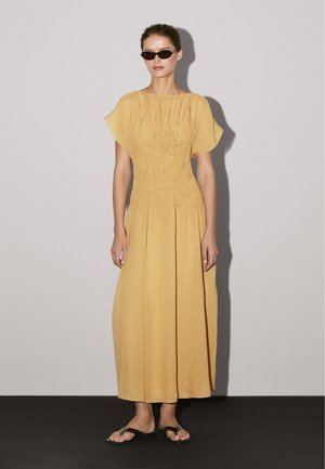 Robe maxi jaune avec de courtes manches volantées, un corsage froncé et une jupe fluide. Associée à des lunettes de soleil noires et des sandales plates.