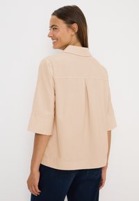 Mujer sonriendo por encima del hombro, vistiendo una camisa beige de manga corta con cuello y unos vaqueros azul oscuro, frente a un fondo blanco liso.