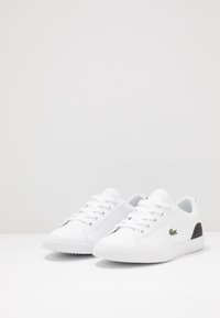 Valkoiset nahkaiset tennarit, joissa on sileä pinta, pyöreä kärki, klassiset nauhat ja musta yksityiskohta kantapäässä. Lacoste-logo näkyvissä.