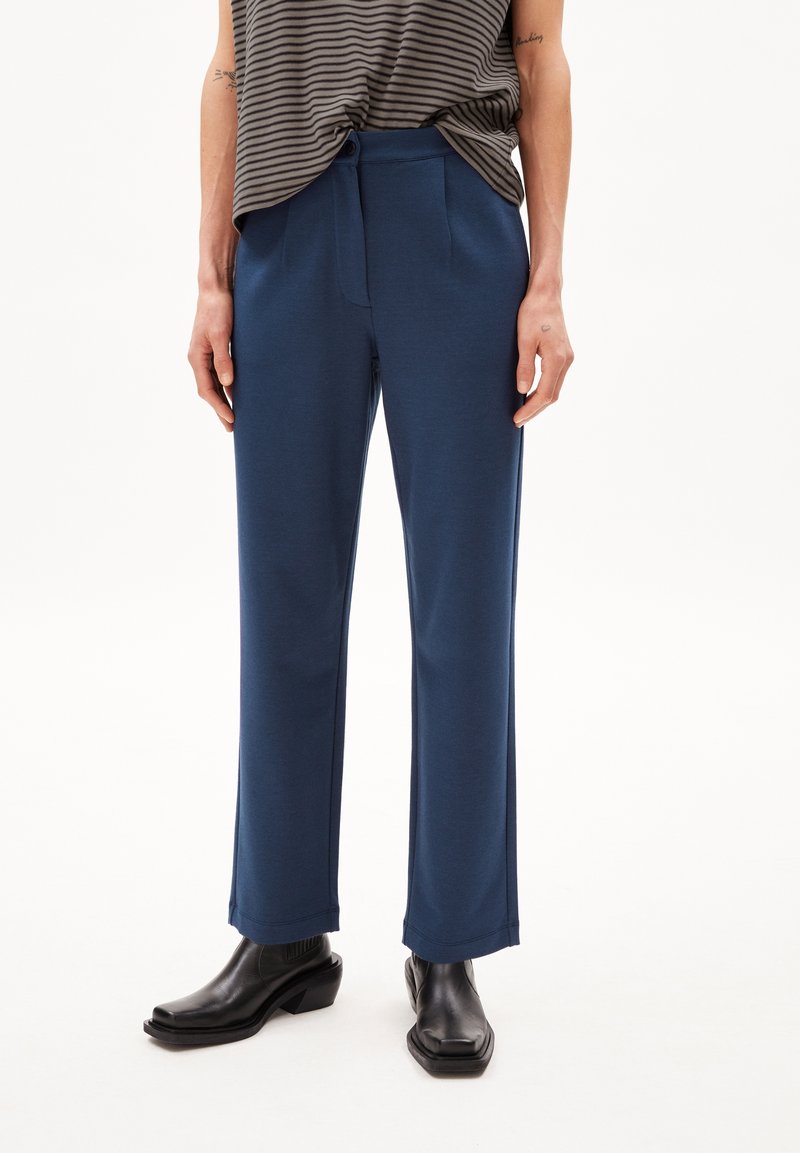 Pantalon bleu marine à jambes larges en tissu doux, présentant une texture lisse, des plis avant et une ceinture cintrée. Porté avec des bottines noires à talons.