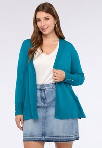 Kobieta w turkusowym kardiganie z złotymi guzikami na białej bluzce oraz w wyblakłych dżinsowych spódnicy z postrzępionym dołem, uśmiechająca się do kamery.
