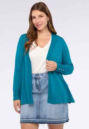 Donna che indossa un cardigan turchese con bottoni dorati sopra una maglietta bianca e una gonna di jeans scolorita con orlo sfrangiato, mentre sorride verso la fotocamera.