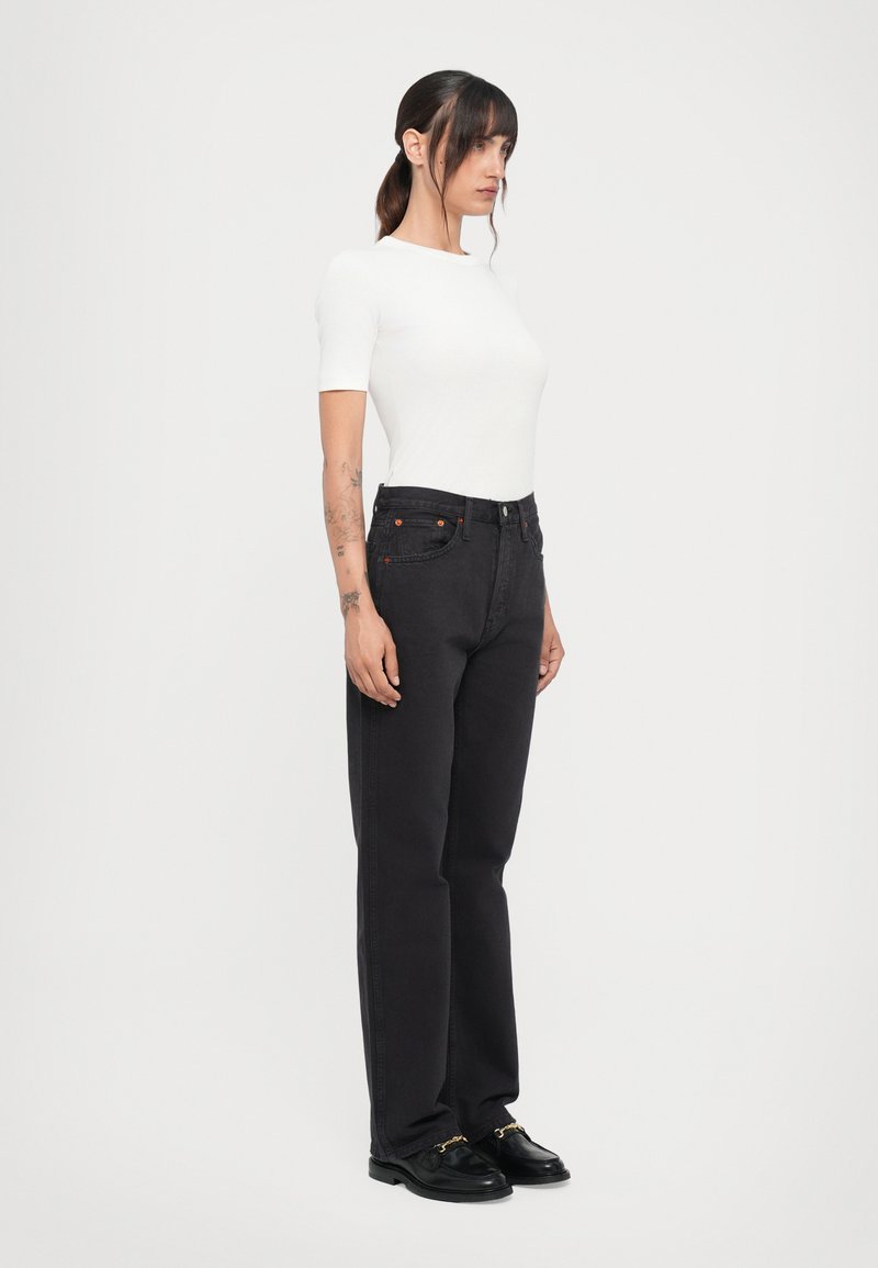 Jean noir taille haute avec jambes droites, accents cousus, cinq poches et un bouton orange. Associé à un haut ajusté à manches courtes blanc.