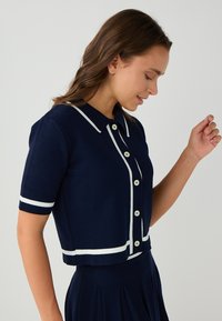 Marineblauer Kurzarm-Cardigan mit weißem Rand und Knöpfen, der einen Kragenhals und eine taillierte Silhouette aufweist. Strukturierter Strickstoff.