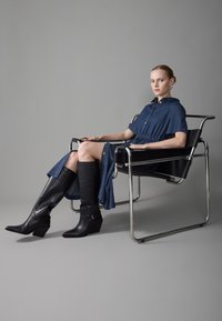 Denim skjortekjole med korte ermer, med knapdetaljer, sammen med høye svarte lærwedge-støvler. Plassert mot en grå bakgrunn.