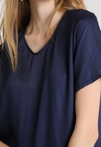 Kvinna iklädd en marinblå V-ringad kortärmad topp, med ljust brunt hår som faller över axeln.