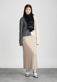 Modèle portant un pull en laine torsadée gris, un cardigan crème, une jupe beige, une écharpe noire Moschino avec un détail en cœur, et des sandales noir et blanc.