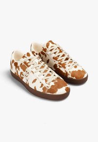 Zapatillas con patrón de piel de vaca, con una base beige y manchas marrones, que cuentan con cordones planos beige y una suela de goma marrón. Textura suave.