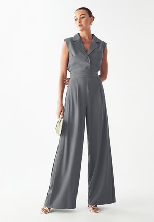 Grauer ärmelloser Jumpsuit mit einem Reverskragen, Knopfverschluss, tailliertem Schnitt und weitem Bein. Das Modell hält eine kleine weiße Handtasche.