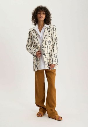 Femme aux cheveux bouclés portant un blazer blanc à motifs floraux, une chemise blanche, un pantalon marron à jambes larges et des sandales beige, debout devant un fond uni.