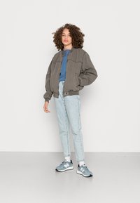 Jaqueta bomber cinza com punhos canelados, blusa azul por baixo, jeans azul claro e ténis cinzentos com solas brancas. Fundo plain.