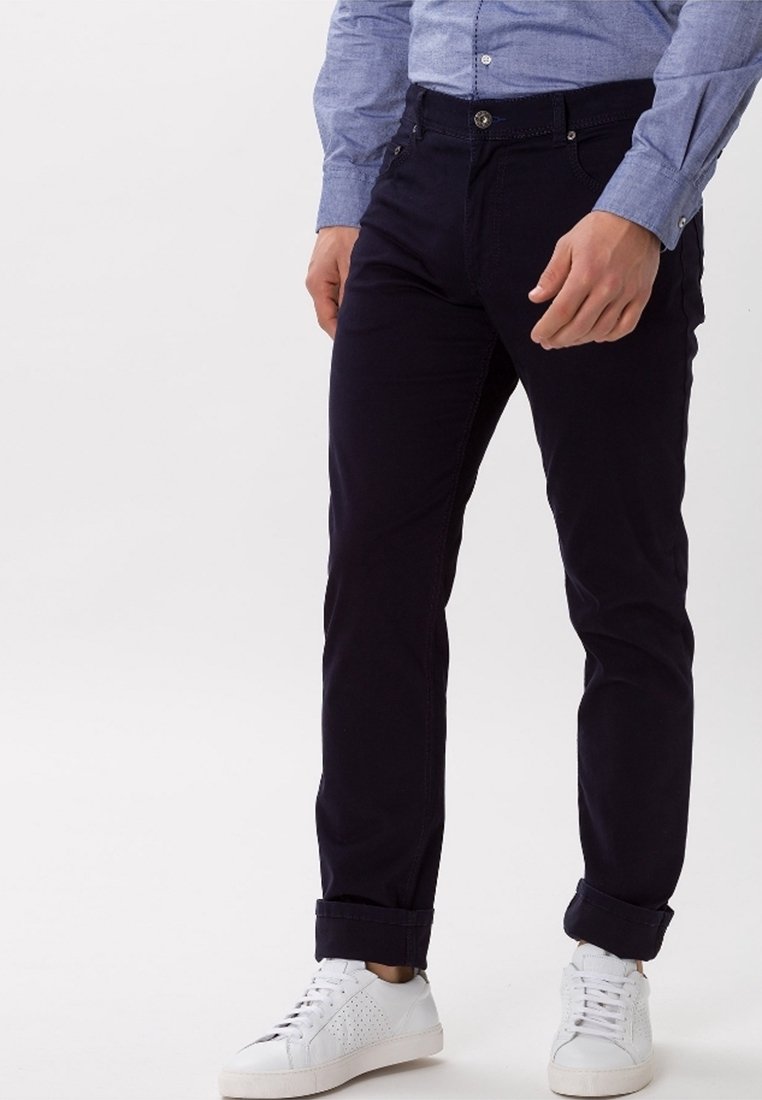 Homme portant un pantalon bleu foncé retroussé aux chevilles, une chemise bleu clair boutonnée et des baskets blanches, debout devant un fond blanc.