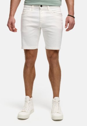 Homme portant un short en jean blanc et des baskets montantes blanches, debout devant un fond uni.