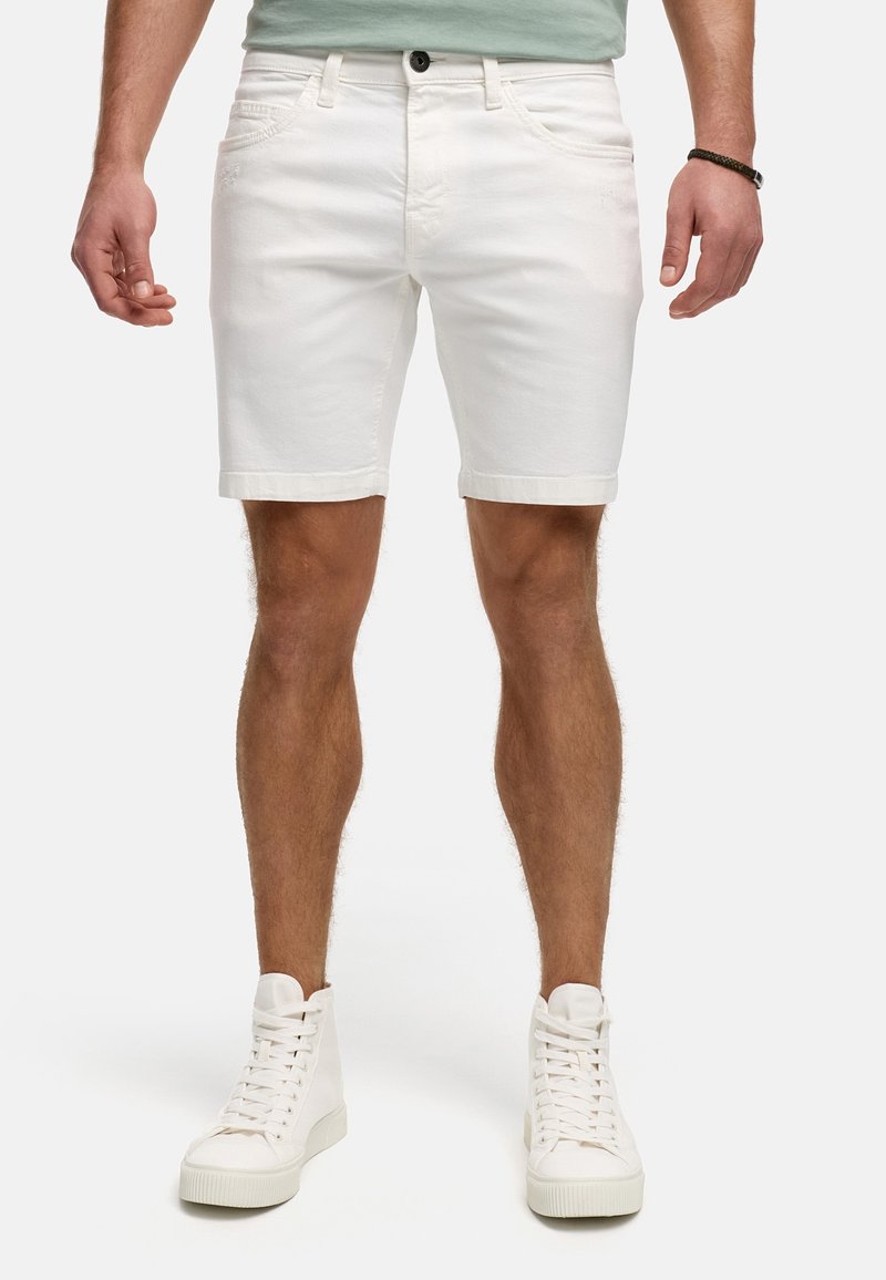 Homme portant un short en jean blanc et des baskets montantes blanches, debout devant un fond uni.