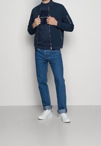 Giacca bomber blu navy con zip sopra una maglietta scura con la scritta "ORIGIN", abbinata a jeans azzurri arrotolati e sneakers bianche.