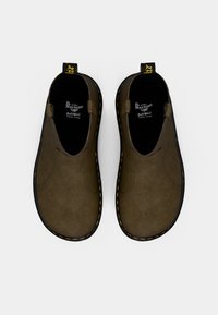 Brązowe zamszowe buty slip-on o gładkiej teksturze, czarna gumowa podeszwa i żółte przeszycia. Zawierają pętelkę na pięcie ułatwiającą zakładanie.