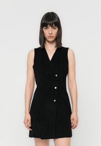 Robe sans manches en velours côtelé noir avec un décolleté en V et des boutons en argent, présentant une silhouette ajustée et un tissu texturé.