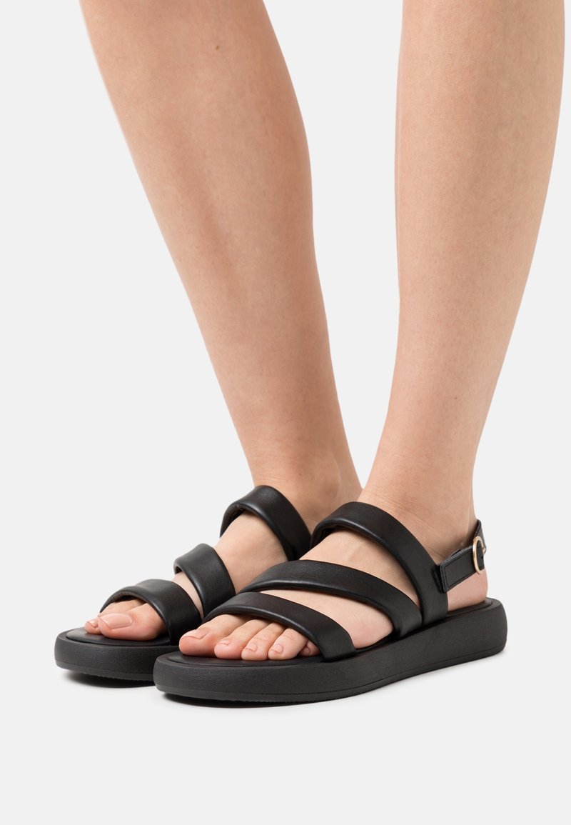 Pieds portant des sandales noires avec trois larges brides et une boucle sur une semelle épaisse, posés sur un fond clair.