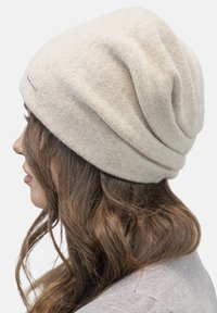 Beige gebreide beanie met een slouchy ontwerp, zachte textuur en een klein metalen label aan de zijkant, geschikt voor koud weer.