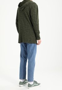 Felpa verde a maniche lunghe con finitura testurizzata, abbinata a jeans dritti blu chiaro e sneaker verdi con suole bianche.