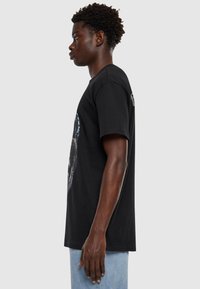 Černé bavlněné tričko s kulatým výstřihem, krátkými rukávy, s grafickým potiskem na přední straně a decentním logem na zadní straně.