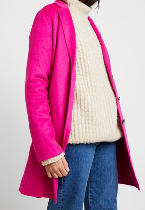 Fuchsia wolmix jas met een getailleerde pasvorm, enkele sluiting en notch-kraag, gelaagd over een crèmekleurige gebreide trui en blauwe jeans.