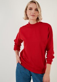 Sudadera roja con cuello alto y mangas largas. Presenta una textura suave y puños acanalados. Lleva puesta sobre pantalones de mezclilla azules.