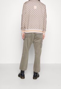 Giacca tessuta con un motivo circolare ripetuto in beige e marrone, con accenti arancioni. Abbinata a pantaloni verde oliva a zampa d'elefante e stivali neri.
